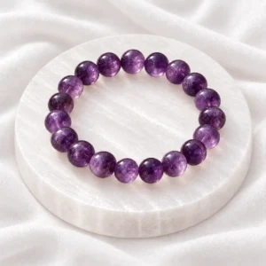 Amethyst Serenity Crystal Bracelet with polished purple crystal beads displayed on a round white selenite plate over soft white fabric.