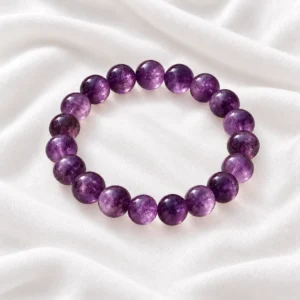 Amethyst Crystal Bracelet with polished purple crystal beads displayed on a round white selenite plate over soft white fabric.