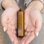 Tiger Eye crystal point held gently in hands, showing a polished golden-brown gemstone with natural chatoyancy and faceted pointed design