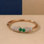 A yellow gold architectural bangle featuring a central emerald-cut green stone and pavé diamonds on a neutral background.