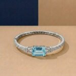 A white gold Art Deco bangle featuring a central emerald-cut blue stone and pavé diamonds on a neutral background.