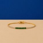 Seven-stone emerald accent lab-grown diamond tennis bracelet in a yellow-toned metal setting on a two-toned background.