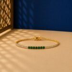 Seven-stone emerald accent lab-grown diamond tennis bracelet in a yellow-toned metal setting on a two-toned background.