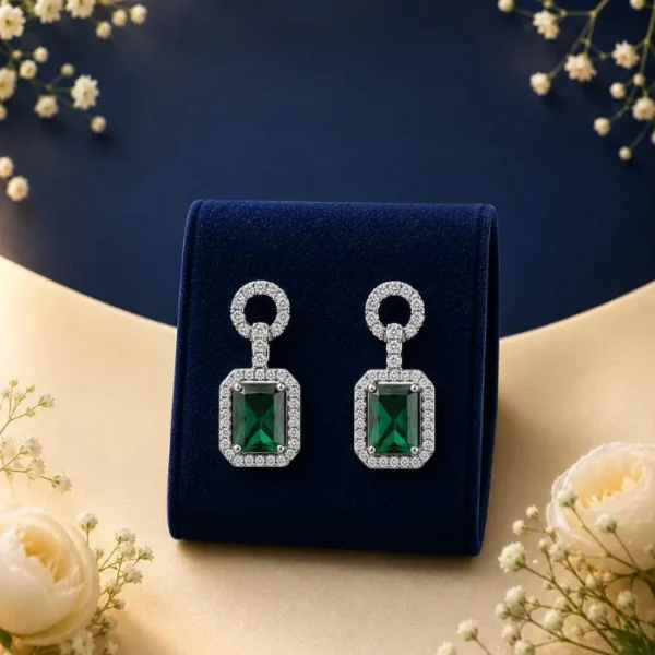 A pair of 18K white gold Art Deco drop earrings featuring emerald-cut green gemstones surrounded by a halo of pave lab-grown diamonds, displayed on a navy blue velvet mount.