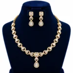 18K Gold Baguette Cut Lab Grown Diamond Halo Necklace Set with Matching Earrings featuring sparkling rectangular baguette diamonds, circular diamond halo links, square diamond centrepiece and elegant pear-shaped drop pendant on a navy blue jewellery display.